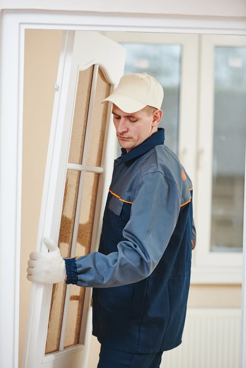 Artisan installateur en tenue de travail bleue et casquette blanche manipule une fenêtre à double vitrage dans un intérieur résidentiel