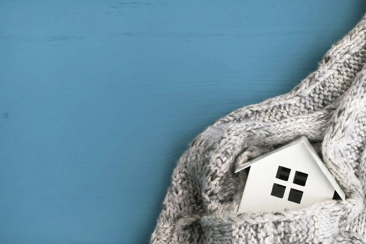 Scène d'intérieur avec maquette de maison blanche enveloppée dans un pull en laine gris chiné à motifs torsadés, sur fond bleu pastel