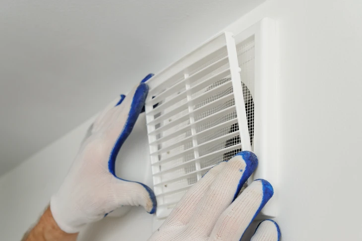 Installateur en gants de protection blancs et bleus positionnant une grille d'aération ventilation dans plafond blanc d'intérieur résidentiel
