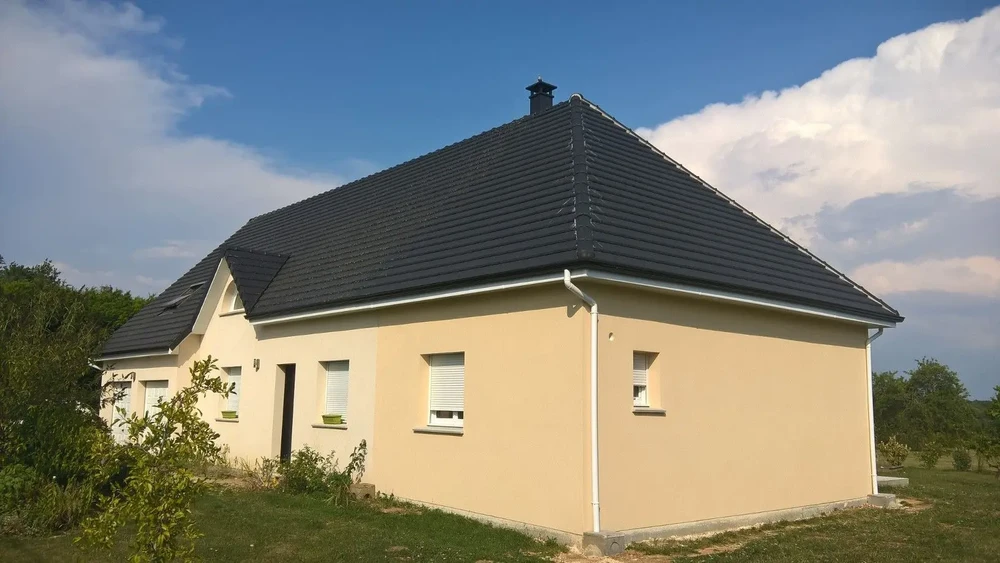 Maison individuelle contemporaine de plain-pied avec façade enduite beige, toiture en tuiles grises à quatre pans, fenêtres à volets roulants blancs et cheminée