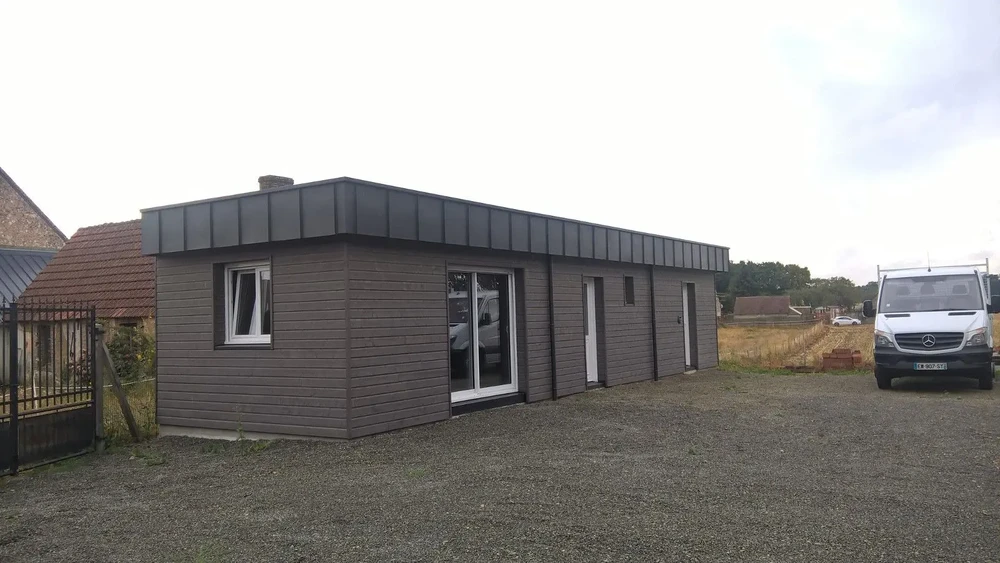 Scène extérieure montrant un bâtiment modulaire neuf en cours de livraison sur terrain de gravier, avec camion Mercedes et maison traditionnelle en arrière-plan