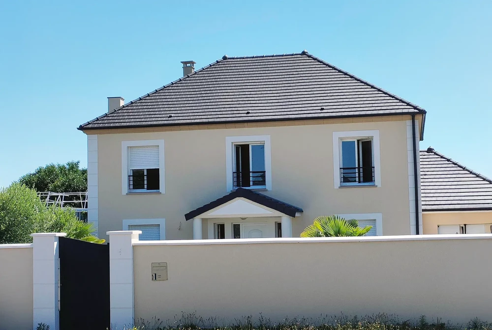 Maison individuelle contemporaine à étage avec façade enduit beige, toiture tuiles grises, trois fenêtres à volets roulants blancs et porte d'entrée sous auvent